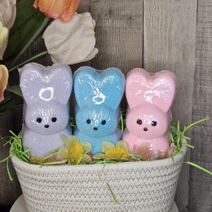 May include: Three bunny-shaped bath bombs in a white woven basket. The bath bombs are pastel colors: lavender, blue, and pink. The basket is decorated with faux grass and small yellow and pink flowers. The background is a wooden panel.