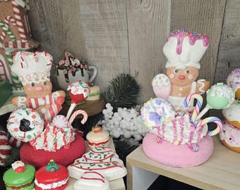 May include: Two Christmas-themed gingerbread figures decorated with candy canes, sprinkles, and frosting. The figures are on a white pedestal with a pink and white donut-shaped base. The figures are surrounded by other Christmas-themed decorations, including a gingerbread house, candy canes, and a Christmas tree.