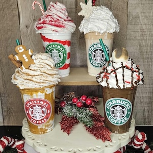 Puede incluir: Cuatro vasos de caf&eacute; Starbucks festivos con crema batida y coberturas navide&ntilde;as. Los vasos est&aacute;n decorados con rayas rojas y blancas, un hombre de jengibre y un copo de nieve. Los vasos est&aacute;n dispuestos en un soporte para pasteles blanco de dos niveles con una cinta roja y blanca.
