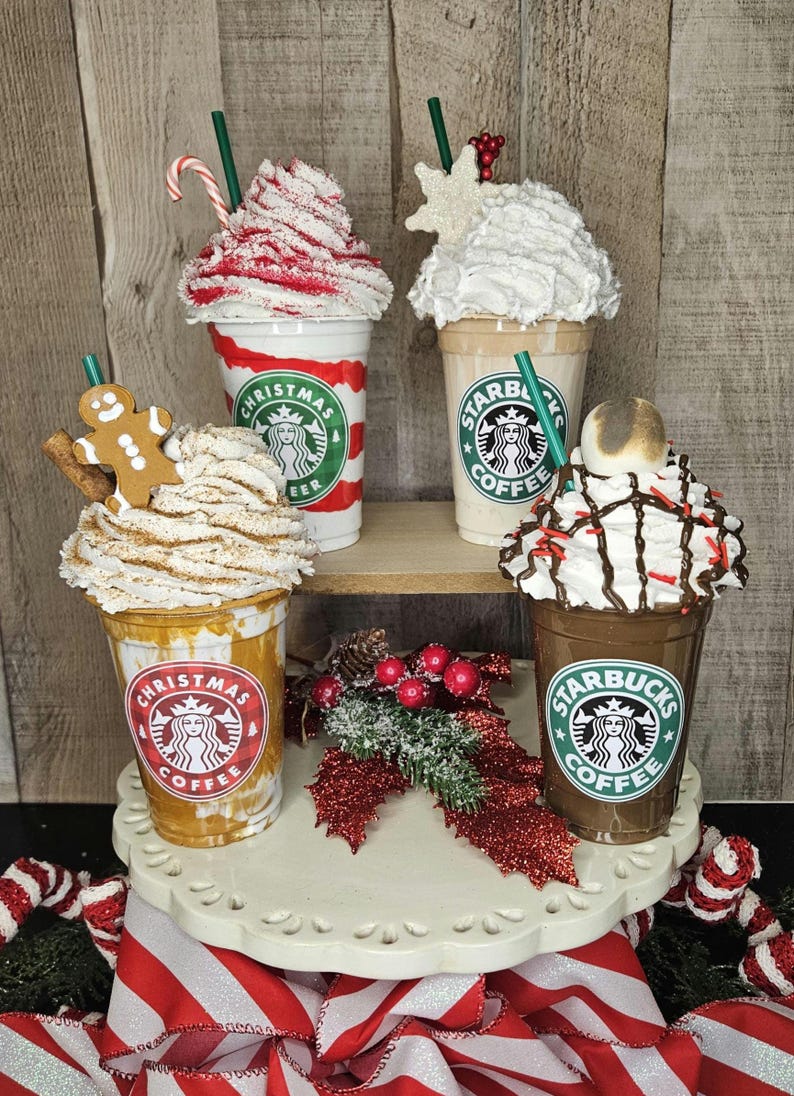 Puede incluir: Cuatro vasos de caf&eacute; Starbucks festivos con crema batida y coberturas navide&ntilde;as. Los vasos est&aacute;n decorados con rayas rojas y blancas, un hombre de jengibre y un copo de nieve. Los vasos est&aacute;n dispuestos en un soporte para pasteles blanco de dos niveles con una cinta roja y blanca.