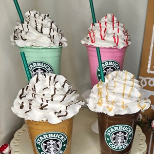 Puede incluir: Cuatro vasos de caf&eacute; Starbucks con crema batida y diferentes ingredientes. Los vasos est&aacute;n dispuestos en fila sobre una superficie blanca. Los vasos son de diferentes colores, incluyendo verde, rosa, marr&oacute;n y beige. Cada vaso tiene un logotipo verde de Starbucks con las palabras "Starbucks Coffee" impresas en &eacute;l.
