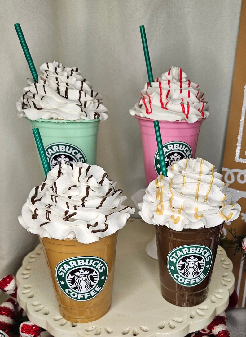 Puede incluir: Cuatro vasos de caf&eacute; Starbucks con crema batida y diferentes ingredientes. Los vasos est&aacute;n dispuestos en fila sobre una superficie blanca. Los vasos son de diferentes colores, incluyendo verde, rosa, marr&oacute;n y beige. Cada vaso tiene un logotipo verde de Starbucks con las palabras "Starbucks Coffee" impresas en &eacute;l.