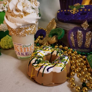 May include: A king cake donut with purple, green, and gold icing and sprinkles. The donut is surrounded by gold beads and a purple and gold mask.