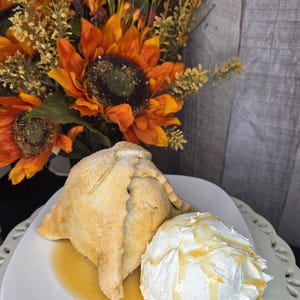 May include: A golden-brown baked pastry with a sugar-dusted crust sits on a white plate next to a large scoop of white ice cream drizzled with amber-colored syrup. The pastry has a triangular, folded shape with visible seams and a textured surface.