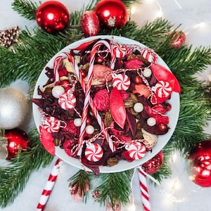 May include: A festive Christmas arrangement featuring a white bowl filled with red and white peppermint candies, dried botanicals, and candy canes. The bowl is surrounded by evergreen branches, red ornaments, and twinkling fairy lights, creating a holiday atmosphere.