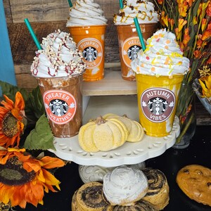 Puede incluir: Cuatro bebidas de caf&eacute; helado con tem&aacute;tica oto&ntilde;al con crema batida y aderezos. Las bebidas est&aacute;n en vasos de pl&aacute;stico transparente con dise&ntilde;os oto&ntilde;ales festivos. Los vasos est&aacute;n dispuestos en un soporte para pasteles blanco de dos niveles con dos bombas de ba&ntilde;o en forma de calabaza amarillas y rollos de canela.