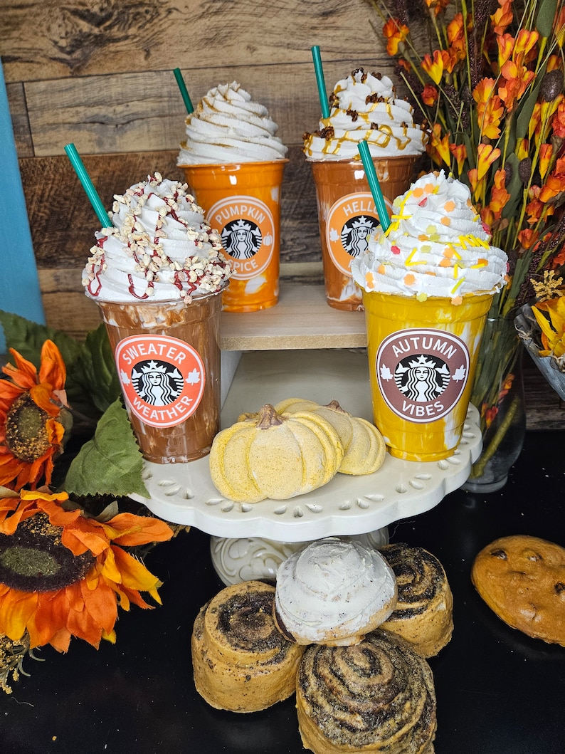 Puede incluir: Cuatro bebidas de caf&eacute; helado con tem&aacute;tica oto&ntilde;al con crema batida y aderezos. Las bebidas est&aacute;n en vasos de pl&aacute;stico transparente con dise&ntilde;os oto&ntilde;ales festivos. Los vasos est&aacute;n dispuestos en un soporte para pasteles blanco de dos niveles con dos bombas de ba&ntilde;o en forma de calabaza amarillas y rollos de canela.