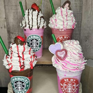 Puede incluir: Cuatro bebidas de caf&eacute; decoradas con crema batida y coberturas. Las bebidas est&aacute;n en vasos de pl&aacute;stico transparente con una base rosa, marr&oacute;n y blanca. Los vasos tienen el logotipo de Starbucks y el texto "Be My Valentine" y "Starbucks Coffee".