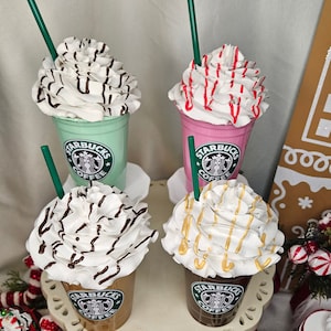 Puede incluir: Cuatro Frappuccinos Starbucks con crema batida y diferentes ingredientes. Los Frappuccinos est&aacute;n en vasos de pl&aacute;stico transparente con tapas verdes y el logotipo de Starbucks. Los ingredientes incluyen llovizna de chocolate, chispas rojas, llovizna de caramelo y virutas de chocolate.