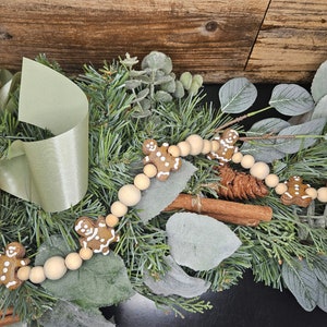 May include: A garland of wooden beads with gingerbread men ornaments. The garland is decorated with green pine branches and cinnamon sticks.