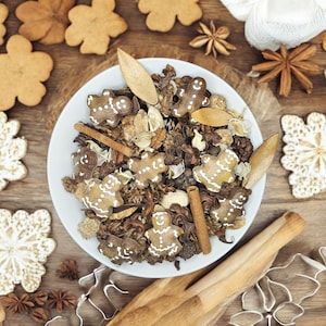 May include: Overhead shot of a white bowl filled with gingerbread men cookies, cinnamon sticks, and dried spices. The bowl sits on a wooden surface surrounded by more cookies, star anise, and cookie cutters, creating a festive holiday scene.