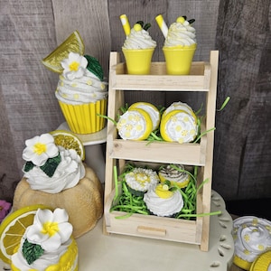 May include: A wooden tiered stand with yellow and white lemon-themed cupcakes and macarons. The cupcakes have white frosting and lemon slices. The macarons have white frosting with yellow lemon designs. The stand is on a white surface with a white lace trim.