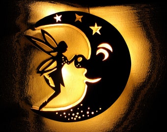 May include: A wooden wall decor featuring a crescent moon with a fairy silhouette inside. The moon has a face and is illuminated from behind, casting a warm glow. The fairy has wings and is holding a flower. The moon is decorated with small stars.