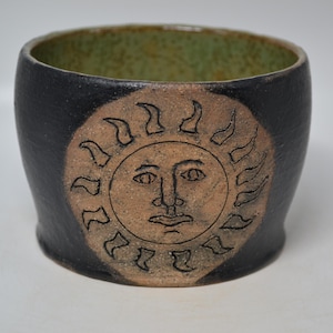 May include: A cylindrical ceramic bowl with a dark charcoal exterior and a textured light green interior. The front features a circular tan medallion embossed with a detailed sun face design, characterized by radiating flame-like rays.