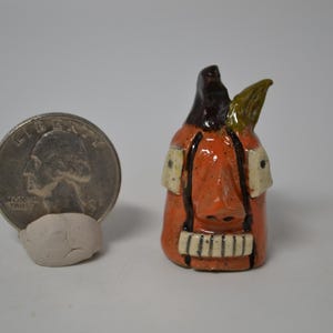May include: A small, orange ceramic figurine with a smiling face, black hair, and a green leaf. The figurine is next to a US quarter coin, providing a size reference. The figurine has a unique, handcrafted appearance.