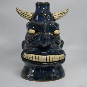 May include: A ceramic tiki mug featuring a dark blue glazed finish with a glossy texture. The mug is shaped like a horned creature with a wide, toothy grin and cream-coloured eyes and horns. The design includes a narrow opening at the top.