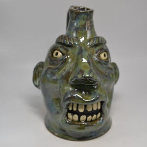 May include: A ceramic face jug with a glossy, mottled green and blue glaze. The vessel features a sculpted human-like face with an aggressive expression, prominent brow, wide nose, and an open mouth revealing a row of individual white teeth.