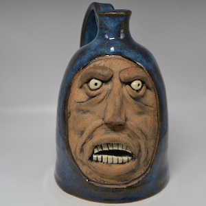 May include: A ceramic jug with a handle, featuring a sculpted face with an angry expression. The jug is primarily blue with a tan face, showcasing detailed facial features like teeth and furrowed brows. The pottery has a glossy finish.