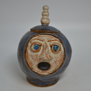 May include: A round ceramic jar with a lid featuring a stacked bead finial. The exterior is a deep blue glaze, while the face is a speckled beige with blue eyes and an open mouth. The vessel has a glossy finish and a sculpted human-like face.