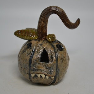 May include: A hand-sculpted ceramic pumpkin figure with a textured tan surface, dark brown vertical grooves, and a curved brown stem. The pumpkin features carved triangular eyes, a jagged mouth with visible white teeth, and a small green leaf.
