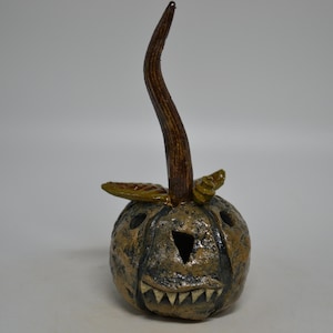 May include: A ceramic pumpkin sculpture with a textured, mottled brown and black finish. It features a tall, curved brown stem, two small green leaves at the base of the stem, a triangular eye cutout, and a wide mouth with visible white teeth.