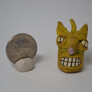 May include: A handmade yellow ceramic sculpture of a creature with a toothy grin, large eyes, and a pink nose. A quarter coin is next to the sculpture for size comparison. The sculpture is on a white background.