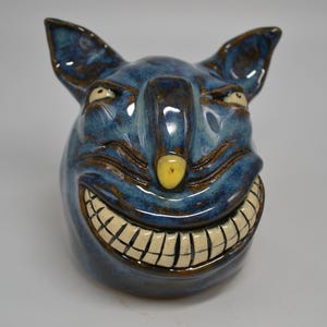 May include: A ceramic sculpture of a grinning, blue creature with large ears and a yellow nose. The sculpture has a glossy finish and a wide, toothy grin. The creature's eyes are small and angled, giving it a mischievous look.