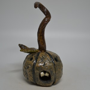 May include: A textured, tan-colored ceramic pumpkin sculpture with a tall, curved brown stem and a single green leaf. The pumpkin features a carved, open mouth revealing two white teeth, along with two circular eye cutouts and dark vertical grooves.