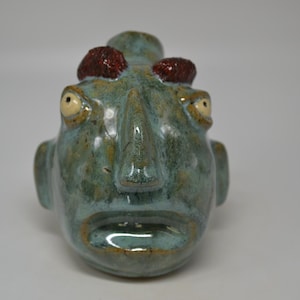 May include: A ceramic sculpture of a face with large, wide eyes and a prominent nose. The face is teal with brown accents and two reddish-brown protrusions on top. The mouth is open, and the sculpture has a glossy finish.