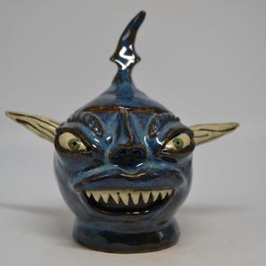 Könnte beinhalten: Ein blau glasiertes Keramikgefäß mit einem Deckel, der ein skulptiertes Kreaturengesicht zeigt. Die Figur hat große grüne Augen, spitze cremefarbene Ohren und einen breiten Mund voller scharfer, weißer Zähne. Oben sitzt ein Flossenaufsatz.