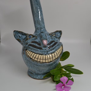 May include: A blue ceramic sculpture of a grinning cat face with a long neck. The cat has a pink nose, blue eyes, and a wide, toothy grin. Green leaves and a pink flower are in the foreground. The sculpture has a speckled texture.