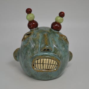 May include: A ceramic sculpture of a face with a teal glaze. The face has a wide grin with visible teeth and two sets of stacked, spherical protrusions on top. The top spheres are red and green.