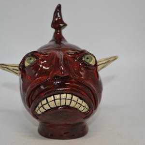 May include: A glossy dark red ceramic sculpture shaped like a creature head with a pointed top protrusion, large green eyes, and a wide mouth displaying a row of white teeth. The figure features two long, pointed, off-white ears extending outward.