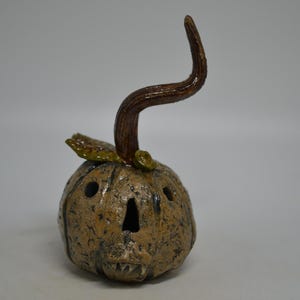 May include: A ceramic pumpkin sculpture with a textured tan and brown finish. It features a long, curved brown stem, a small green leaf, and cutouts for eyes and a mouth with visible teeth, creating a hollow, carved gourd appearance.