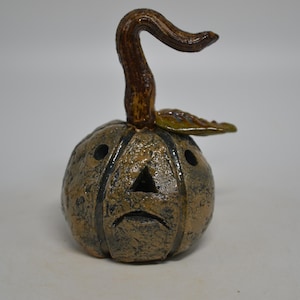 May include: A ceramic pumpkin sculpture with a textured, mottled brown and black finish. It features a curved brown stem, a single green leaf, and carved triangular eyes and a downturned mouth, creating a hollow, lantern-like appearance.