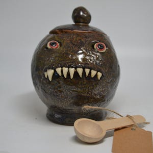 May include: A round, dark brown ceramic jar with a textured surface, featuring two realistic eyes and a row of sharp, white teeth. The jar has a matching lid with a knob handle and is accompanied by a small wooden scoop tied with twine.