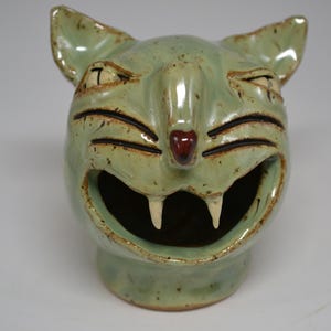 Peut inclure: Un récipient en céramique en forme de chat, de couleur vert clair. Le chat a un large sourire édenté, des oreilles pointues et un petit nez rouge. Le récipient est probablement fait à la main et a un design unique et fantaisiste.