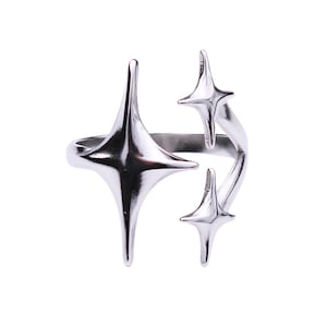 May include: A silver-colored ring featuring a unique design with three star-shaped embellishments. The stars have pointed tips and are arranged in a dynamic, open configuration. The ring is set against a plain white background.