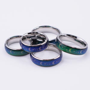 May include: Five stainless steel mood rings. The rings change color from blue to green, with a crescent moon and star design. The rings are displayed on a white background.
