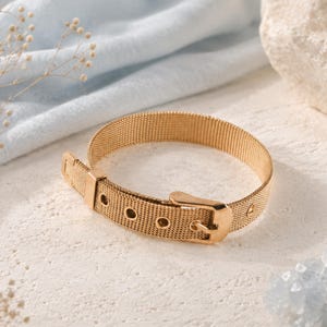 May include: A gold-toned metal mesh bracelet featuring a buckle closure design. The bracelet has a fine, woven texture and includes a metal loop and a series of circular holes along the strap, mimicking the appearance of a traditional belt.