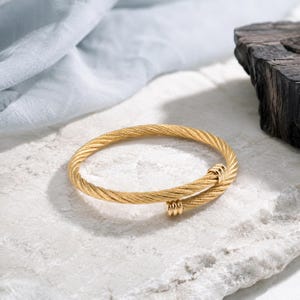 May include: A gold-toned bangle bracelet featuring a twisted cable design. The bracelet has an open-ended style with two small, smooth, cylindrical gold-toned accents at each terminal end, creating a continuous, textured, circular appearance.