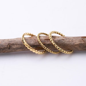 May include: Three gold-toned rings are displayed on a textured brown wooden branch. The first ring features a beaded design, the second ring has a twisted rope texture, and the third ring is composed of a series of small, smooth, connected spheres.