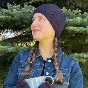 May include: A dark purple knit beanie with a cable knit pattern, worn with a blue denim jacket and dark purple knit gloves, while holding a light blue ceramic mug. The beanie features a textured, braided design and a snug fit around the head.