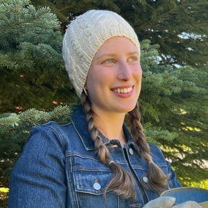 May include: A cream-colored knit beanie with a textured cable pattern, worn with a dark blue denim jacket featuring silver-toned metal buttons and a visible chest pocket, paired with a blue ceramic mug held in the hands.