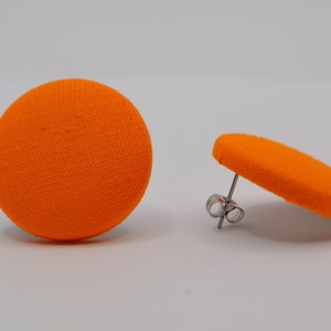 Könnte beinhalten: Ein Paar leuchtend orangefarbene Ohrstecker mit Stoffbezug. Ein Ohrring steht aufrecht und zeigt den Stecker und die Rückseite, während der andere flach liegt. Die Ohrringe haben eine runde Form und eine strukturierte Oberfläche.