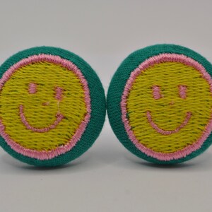 Peut inclure: Deux boutons en tissu turquoise avec des coutures roses autour des bords. Chaque bouton a un smiley jaune brodé au centre.