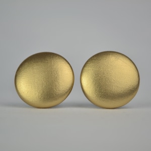May include: A pair of gold-coloured, circular buttons with a textured pattern. The buttons are of equal size and shape, set against a plain background. These buttons could be used for clothing or craft purposes.