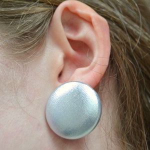 Könnte beinhalten: Ein großer, runder silberner Ohrring mit einer strukturierten Stoffoberfläche. Der Ohrring befindet sich in der Nähe des Ohrs und zeigt seine Größe und sein Design. Der metallische Glanz des Ohrrings deutet auf einen modernen, minimalistischen Stil hin.