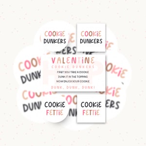 May include: A collection of circular and square stickers featuring the text COOKIE DUNKERS and COOKIE FETTIE in pink, brown, and black. A rectangular card displays VALENTINE COOKIE DUNKERS with instructions: FIRST YOU TAKE A COOKIE, DUNK IT IN THE TOPPING, NOW ENJOY YOUR COOKIE, DUNK, DUNK, DUNK!