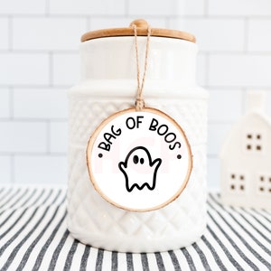 Pu&ograve; includere: Un barattolo di ceramica bianco con coperchio in legno e un'etichetta decorativa con la scritta "BAG OF BOOS" e un'illustrazione di un fantasma. Il barattolo &egrave; su una superficie a righe, con una piccola statuina bianca a forma di casa sullo sfondo.